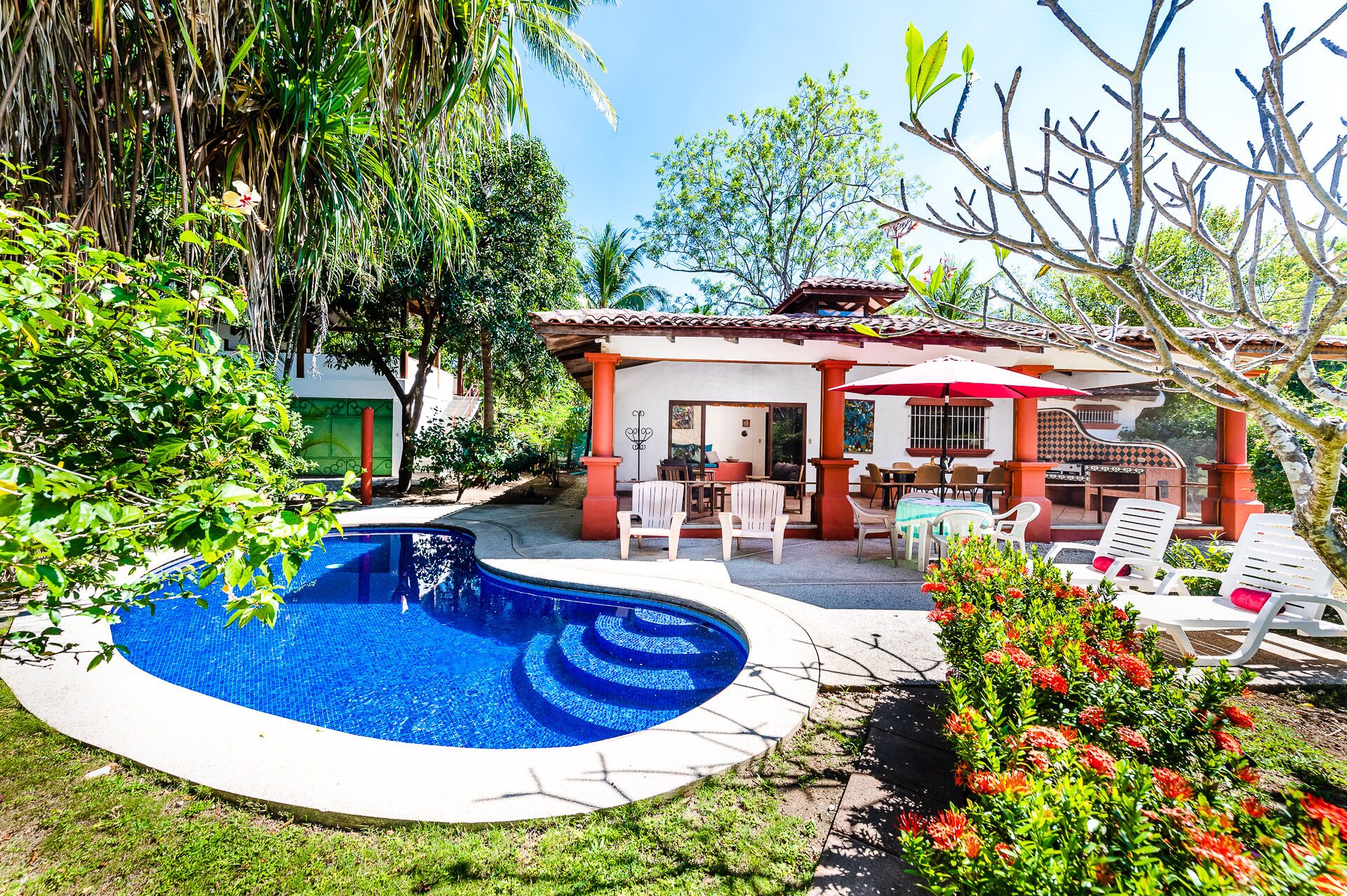 Piscina cercada por um terraço com mobiliário de exterior, área de estar coberta e vegetação exuberante.