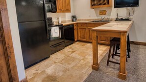 A kitchen with wooden cabinets, a black refrigerator, a microwave, a dishwasher, a wooden table, and a television mounted on the wall. - Queen Suite at Ponds Lodge Cabin 31 Unit 1 (Island Park)