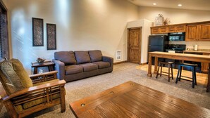 A living room with a sofa, a wooden coffee table, a wooden chair, and a kitchen area with wooden cabinets and appliances. - Queen Suite at Ponds Lodge Cabin 31 Unit 1 (Island Park)