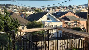 Terraza de madera con barandilla de metal, plantas en macetas y vista a un barrio residencial con casas y cielo despejado.