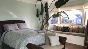 Un dormitorio con una cama, un sofá de mimbre, un asiento junto a la ventana con cojines y una planta grande.