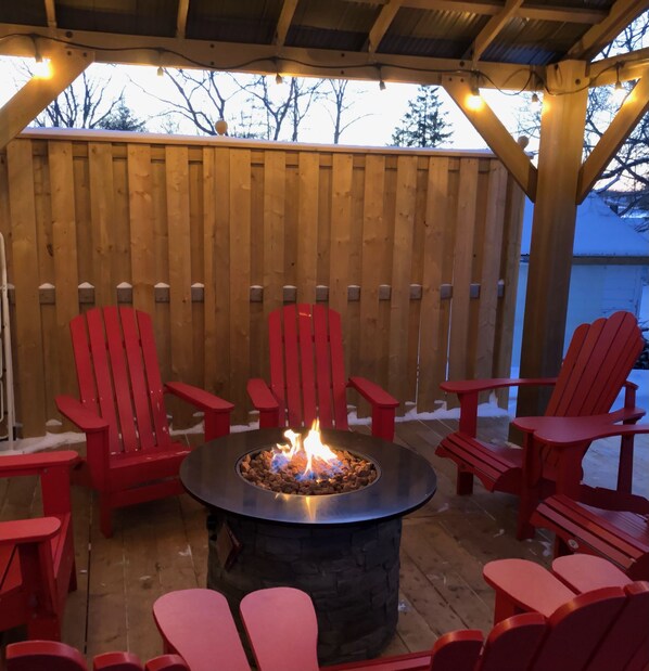 Un foyer circulaire, entouré de chaises rouges, installé sur une terrasse en bois, avec une clôture en bois en arrière-plan.
