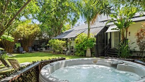 Un bain à remous entouré d’une végétation luxuriante, une terrasse en bois et un bâtiment rustique avec un toit en métal.