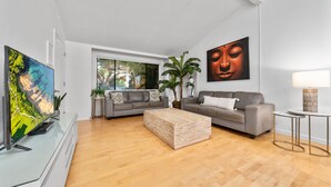 Un salon moderne avec un canapé gris, une table basse en bois, un téléviseur posé sur un meuble en verre, une grande fenêtre et un tableau accroché au mur.
