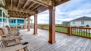 Une terrasse en bois dotée d’un espace salon couvert, avec vue sur une maison et un paysage verdoyant.