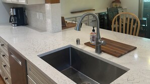 Eine Küche mit weißem Marmorbelag, Spüle aus Edelstahl und Holzschränken. Zwei Pendelleuchten mit gelben Lampenschirmen und ein Holzbalken an der Decke.