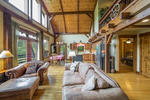 A spacious living room with a high ceiling, wooden beams, a dining area with a table and chairs, and a kitchen with wooden cabinets.