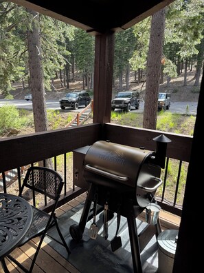 A barbecue grill on a wooden deck with a view of a forest and parked cars.