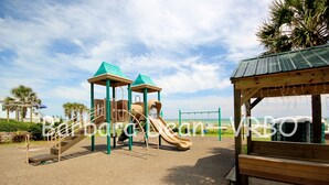 Un parque infantil con tobogán y estructuras para escalar, rodeado de una zona de arena y un refugio de madera.