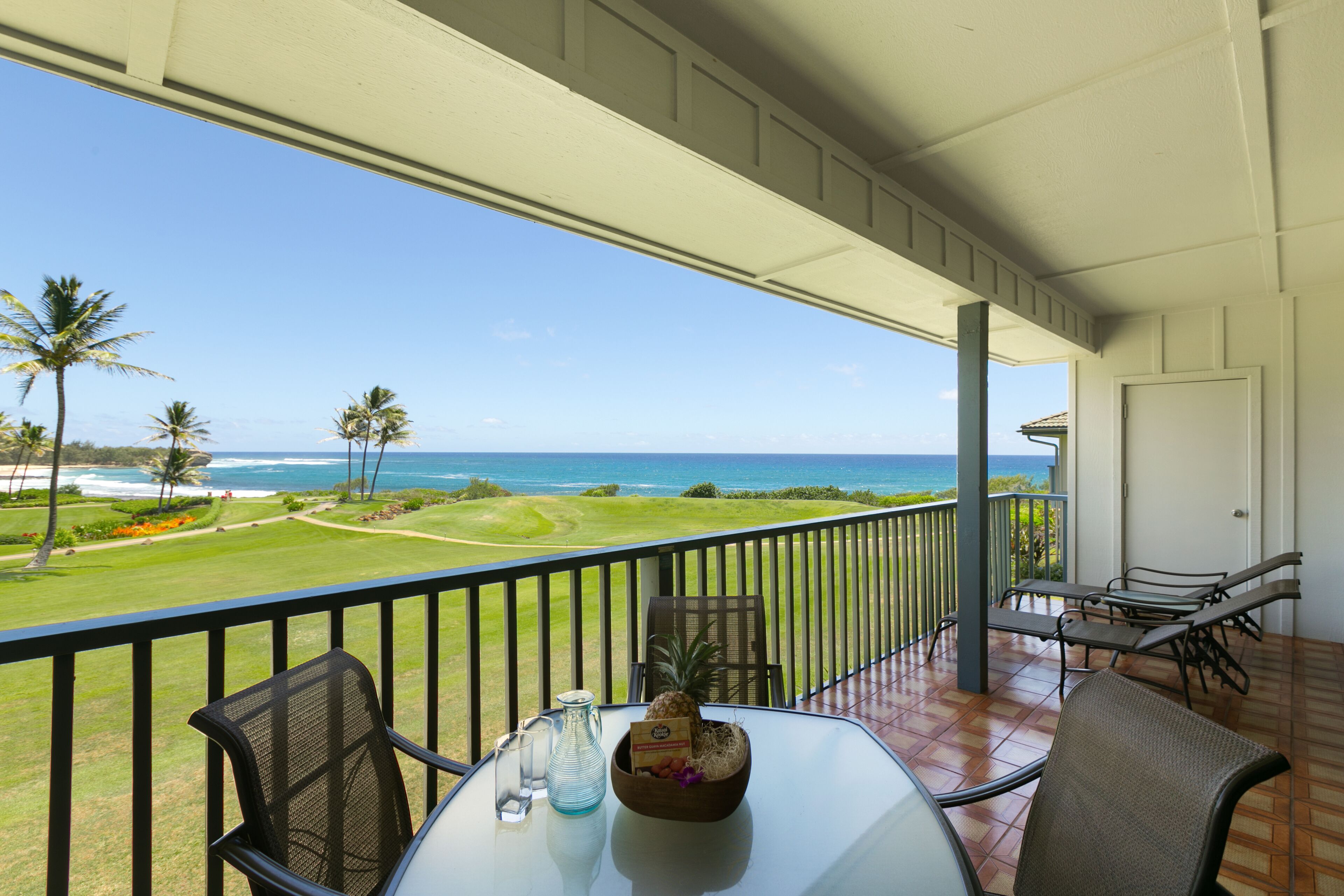 A balcony with a view of the ocean and a golf course. There are chairs and a table with a bottle and a bowl of fruit.