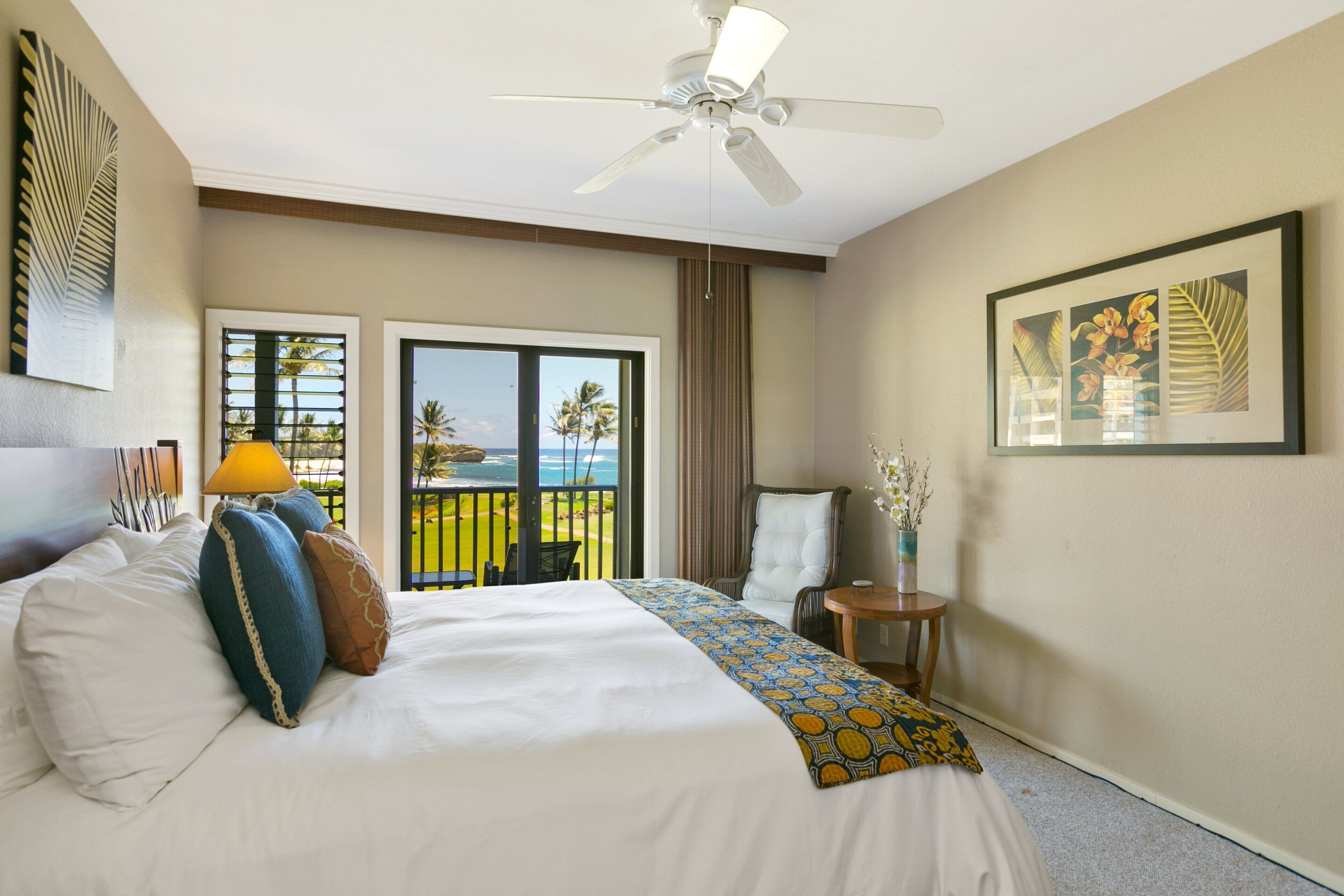 Habitación de hotel con cama, ventilador de techo, balcón con vista a palmeras y un cuadro en la pared.