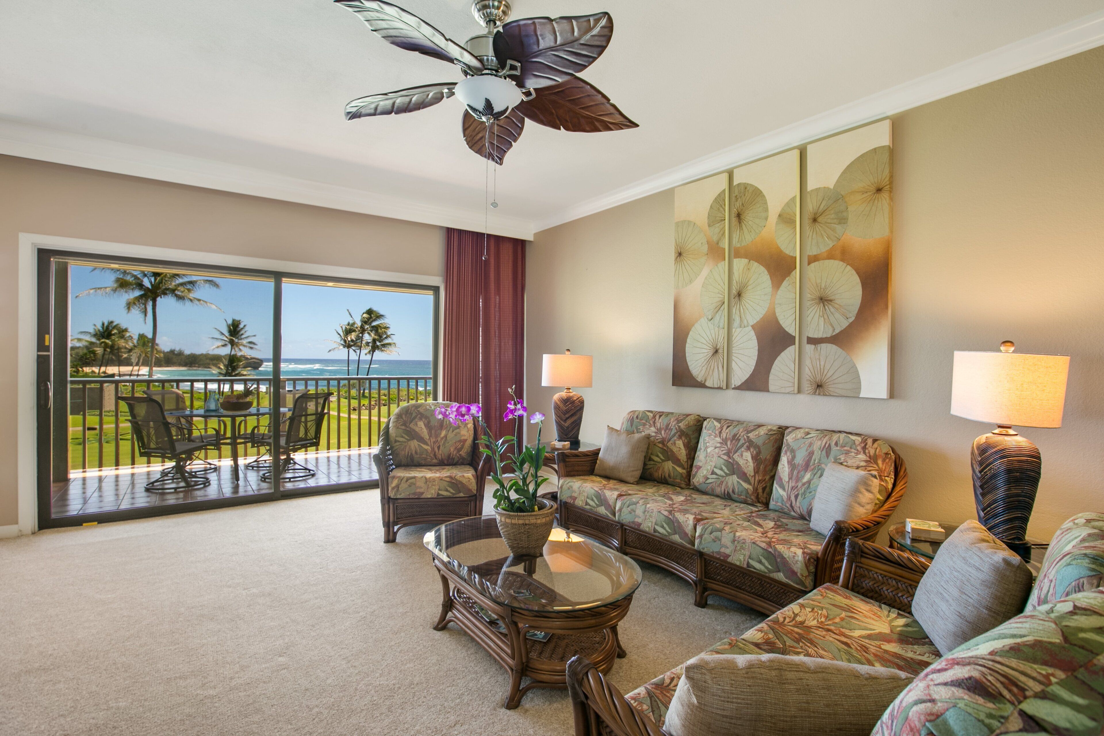 Ein geräumiges Wohnzimmer mit Balkon, Deckenventilator und großem Fenster mit Blick auf Palmen und das Meer.