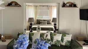 A bedroom with a bed, bedside tables, a TV, and a window with curtains. - Serene Mountain View Condo 11B North Shore Princeville - on Makai Golf Course (Princeville)