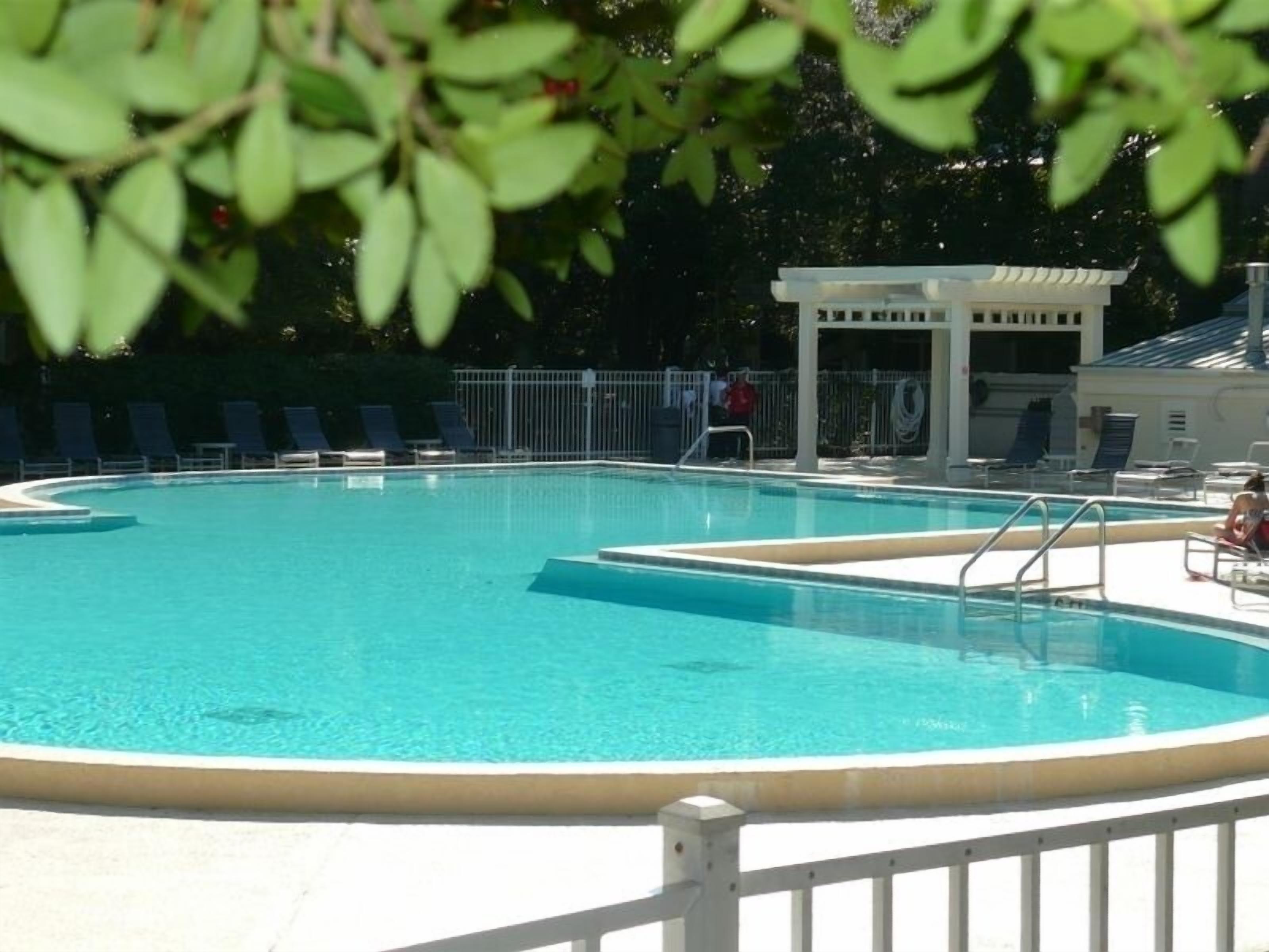 A clear, circular swimming pool with a white deck surrounding it, a covered seating area, and lounge chairs.