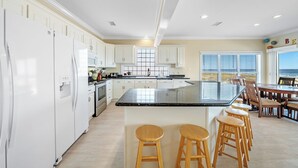 Een ruime keuken met witte kasten, een groot eiland met krukken, en uitzicht op de oceaan door de ramen.