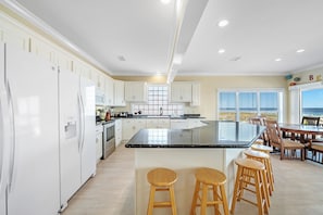 A spacious kitchen with white cabinets, a large island with stools, and a view of the ocean through the windows. - You'll love Seafoam! Beachfront. Pool. 6 BR, sleeps 18. Nice. (Gulf Shores)