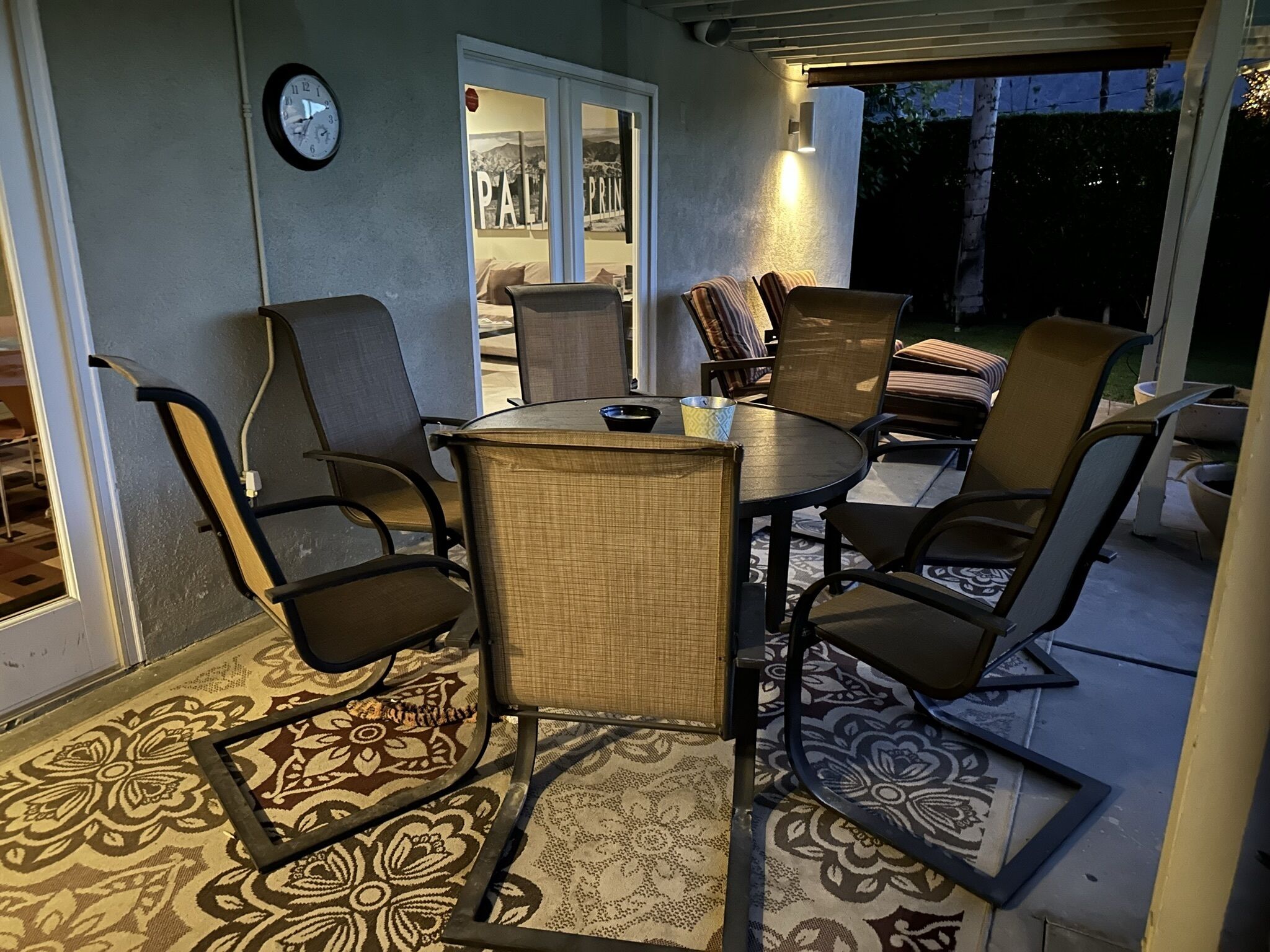 An outdoor patio with a round table and six chairs, a clock on the wall, and a view of the backyard.