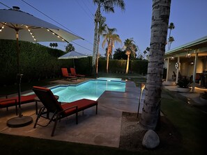 Une piscine entourée de palmiers et d’une haie bien entretenue, avec des chaises longues rouges et des parasols.
