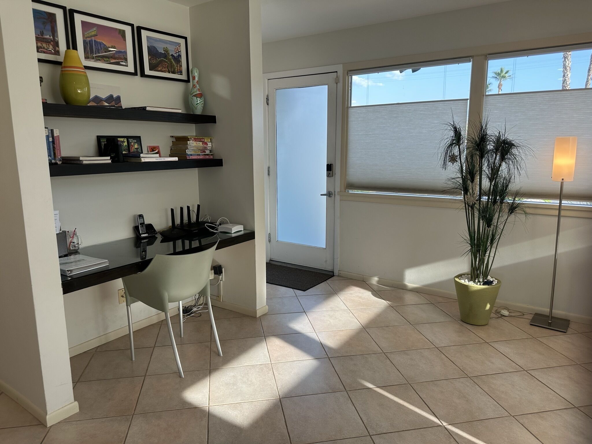 A modern office space with a desk, chair, shelves with books and decorative items, a potted plant, and a window with blinds.
