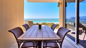 Een balkon met een houten tafel en rieten stoelen, met uitzicht op een strand en een helderblauwe lucht.