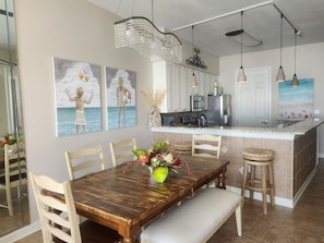 A dining area with a wooden table, chairs, a bench, and a vase with flowers. There are paintings on the wall depicting beach scenes.