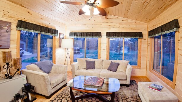 A cozy cabin interior with a wooden ceiling, two sofas, a coffee table, and large windows offering a view of the outdoors.