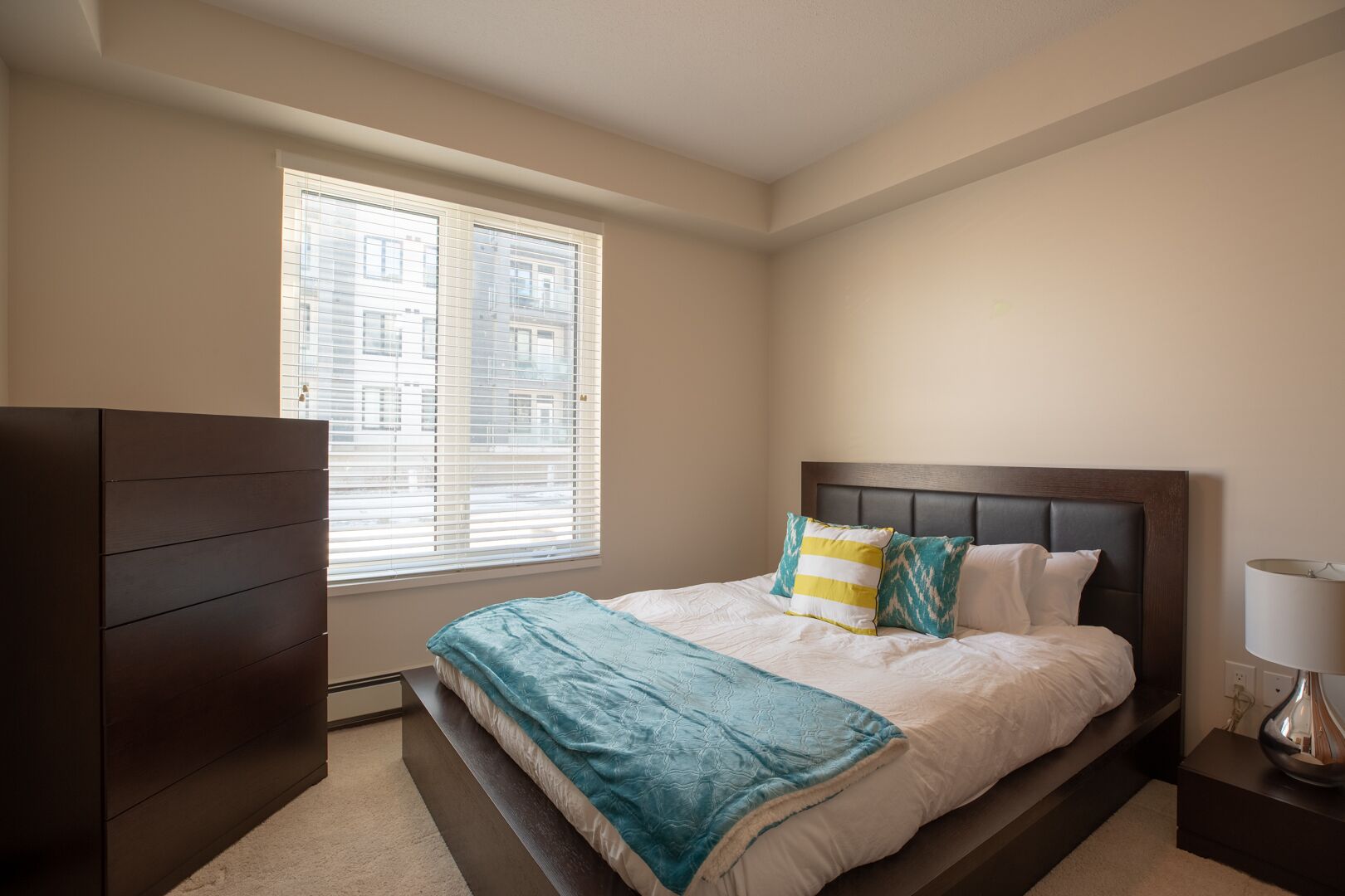 A bedroom with a bed, a dresser, a window with blinds, and a lamp on a bedside table.