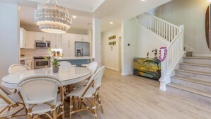 A modern kitchen with white cabinets, a dining area with a glass table and wicker chairs, and a staircase with a white railing. - Leilani Villa: Gardenview Condo on Kauai's North Shore w/ Pool, Spa & AC (Princeville)