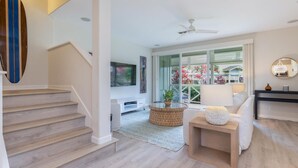 A modern living room with a staircase, a flat-screen TV, a ceiling fan, and a large window with a view of greenery. - Leilani Villa: Gardenview Condo on Kauai's North Shore w/ Pool, Spa & AC (Princeville)