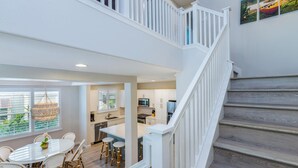 A spacious kitchen with white cabinets, a dining area with a glass table and wicker chairs, and a wooden staircase. - Leilani Villa: Gardenview Condo on Kauai's North Shore w/ Pool, Spa & AC (Princeville)