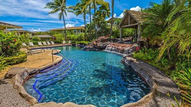 A clear pool with a waterfall feature, surrounded by palm trees and lounge chairs. - Leilani Villa: Gardenview Condo on Kauai's North Shore w/ Pool, Spa & AC (Princeville)