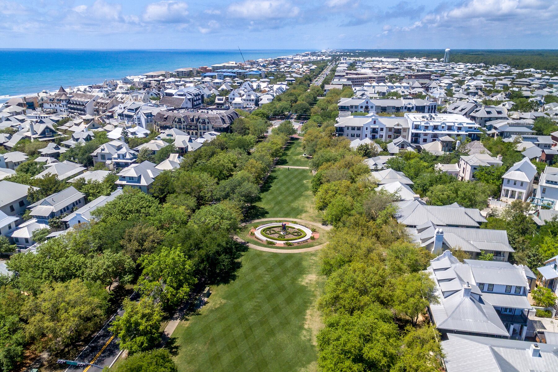 An aerial view of a residential area with houses, a central circular park, and a coastline in the background.