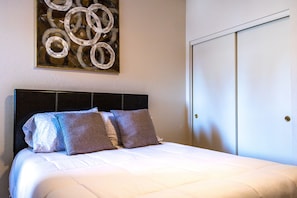 A neatly made bed with a white comforter and two pillows, a dark headboard, and a wall-mounted abstract painting. - Central location, cozy, quiet, gated. Your home base to explore! (Mesa)