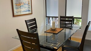 A dining area with a glass table, four wooden chairs, a hanging light fixture, and a framed painting on the wall.