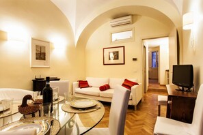 A cozy living room with a round glass table set for dining, a white sofa with red cushions, and a television on a wooden stand.