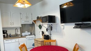 Une cuisine compacte avec des meubles blancs, un four à micro-ondes et une cuisinière. On y trouve une table à manger recouverte d’une nappe rouge et entourée de chaises en bois. Un luminaire est fixé au plafond, au-dessus de la table.