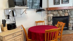 Un coin repas avec une table ronde, des chaises en bois, une nappe rouge, une cheminée en pierre et un téléviseur fixé au mur.