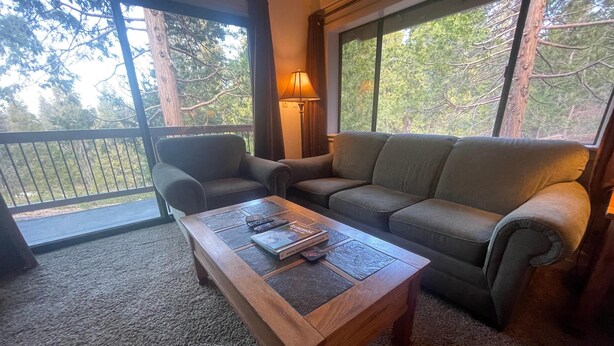 Un salon confortable avec un canapé, une table basse et une lampe. Il y a un balcon avec une balustrade et une vue sur les arbres.