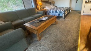 Une pièce comprenant un canapé, un lit recouvert d’une couvre-lit à motifs, une table basse en bois, une lampe et une porte derrière laquelle on aperçoit une machine à café.