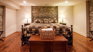 A bedroom with a stone accent wall, a checkered bedspread, two bedside lamps, a wicker basket on the nightstand, and a wooden bench in the center. - 1870's Luxury Guest Cottage, Historic Downtown St. Charles, Mo. (St. Charles)