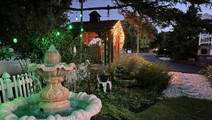 Um jardim com uma fonte decorativa, cerca branca de piquete e um gazebo com luzes de cordão.