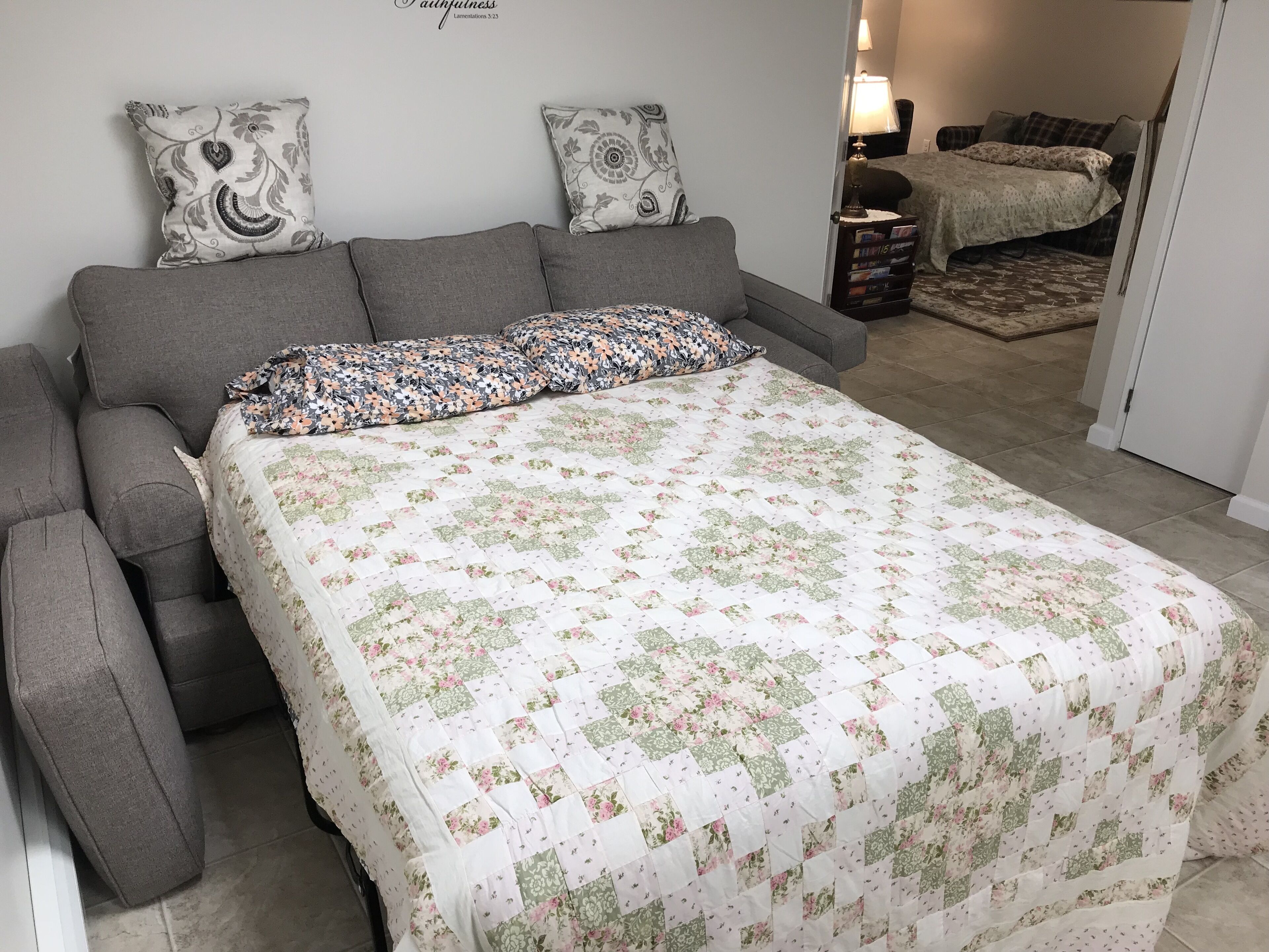 A bedroom with a grey sofa, a floral bedspread, and a nightstand with books.