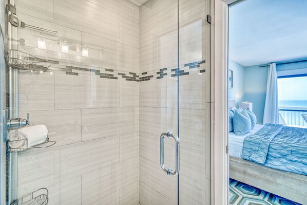 A modern bathroom with a glass shower enclosure, a towel rack, and a visible bed with a blue bedspread in the adjacent room.