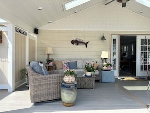 A covered outdoor seating area with wicker furniture, a fish skeleton wall decoration, and a sign reading 'BY THE SEA'.