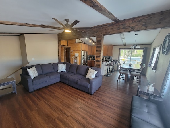 A living room with a sectional sofa, wooden flooring, a ceiling fan, and a kitchen area visible through an open doorway.