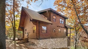A log cabin with a stone foundation, multiple windows, and a sloped roof, surrounded by autumn trees.