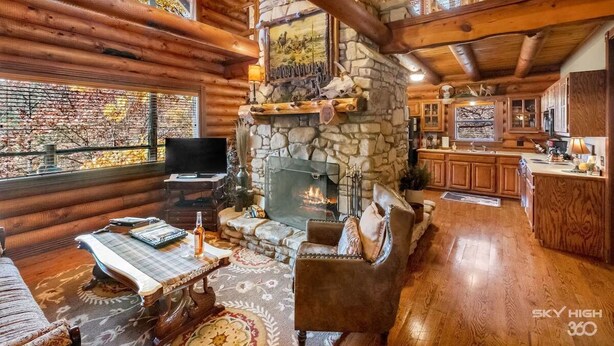 A log cabin interior with a stone fireplace, wooden furniture, and a kitchen area.