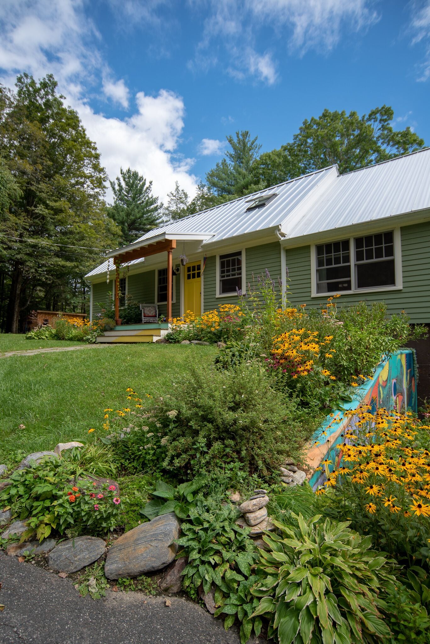 A green house with a porch, surrounded by a garden with various plants and flowers.