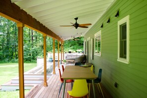 A covered patio with a wooden table and colorful chairs, a ceiling fan, and a view of a green house and forest. - Secluded Family Home in Serene Stockbridge Woods (Stockbridge)