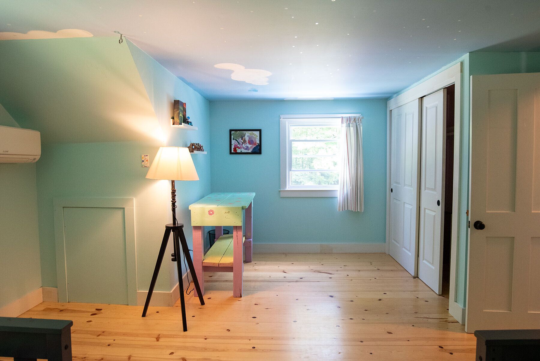 A room with a wooden floor, a desk with a chair, a lamp, a window with curtains, and a door.